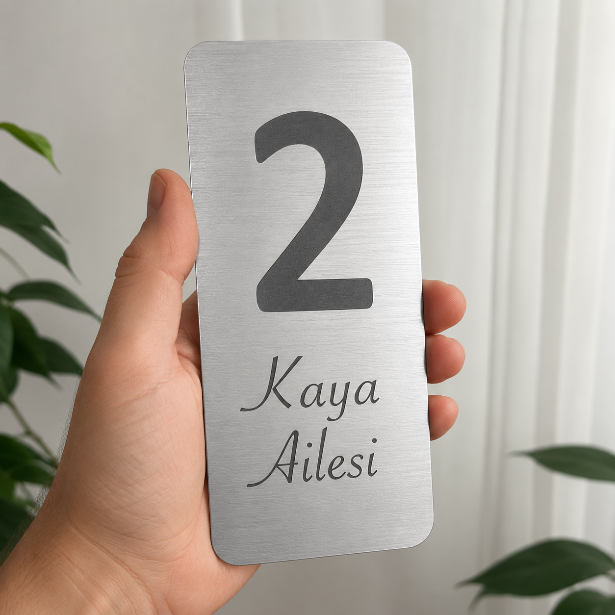 Kişiye özel isimlik kapı tabelası elde tutma görseli, metalik gri pleksi lazer kazıma